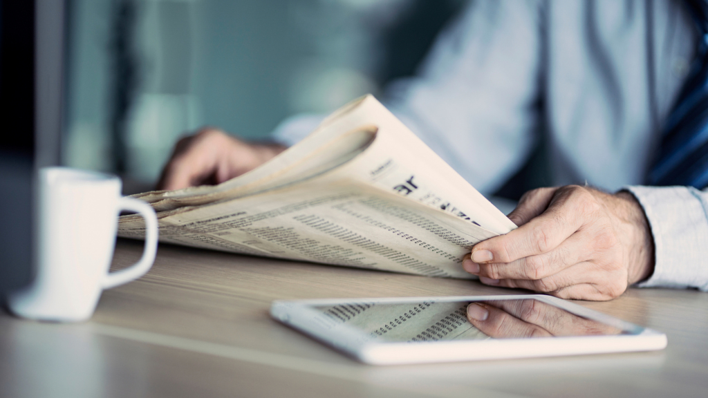 Man’s hands holding newspaper with tablet and coffee mug on table