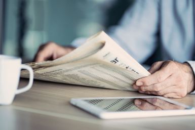 Man’s hands holding newspaper with tablet and coffee mug on table